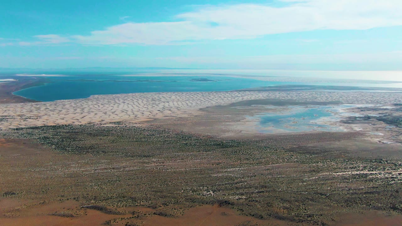 Aerial frame in Guerrero Negro showing turquoise water at the horizon, light tidal flats across the middle, and darker scrub vegetation in the foreground, no visible people.