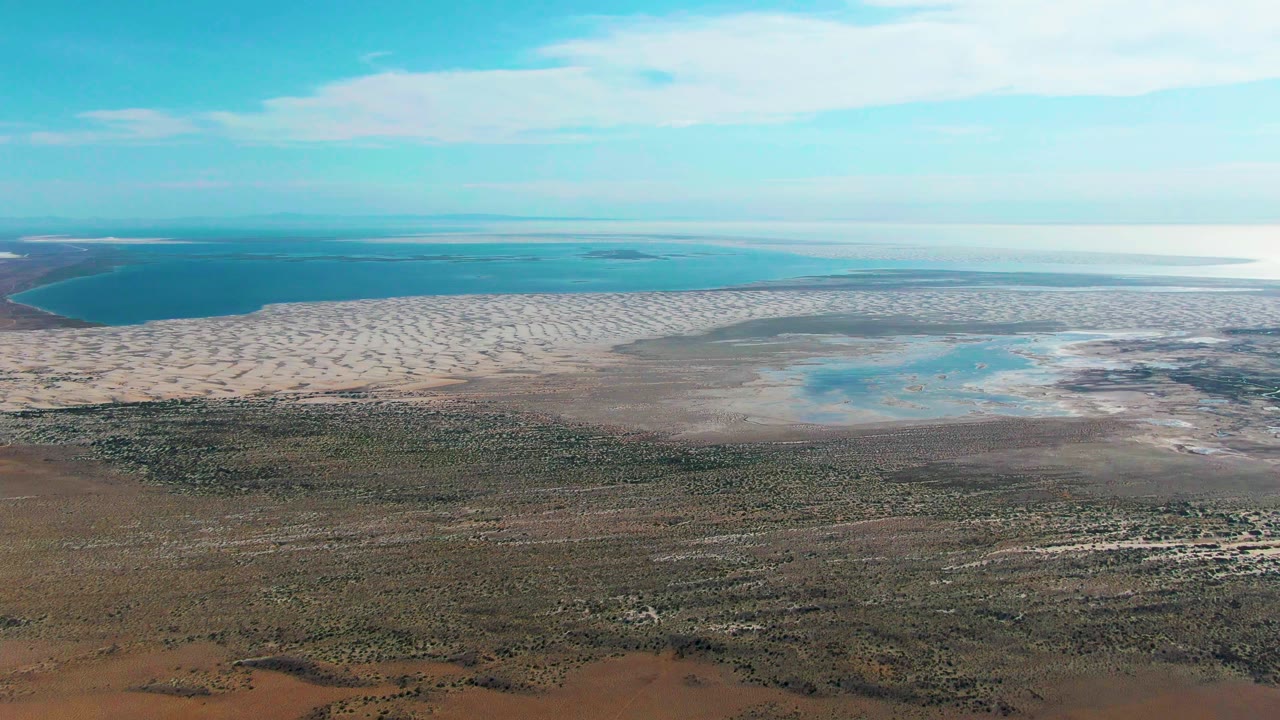 Mid-clip aerial frame over Guerrero Negro with a wide band of pale tidal flats, blue water behind it, and textured brown scrubland in front, no visible people.