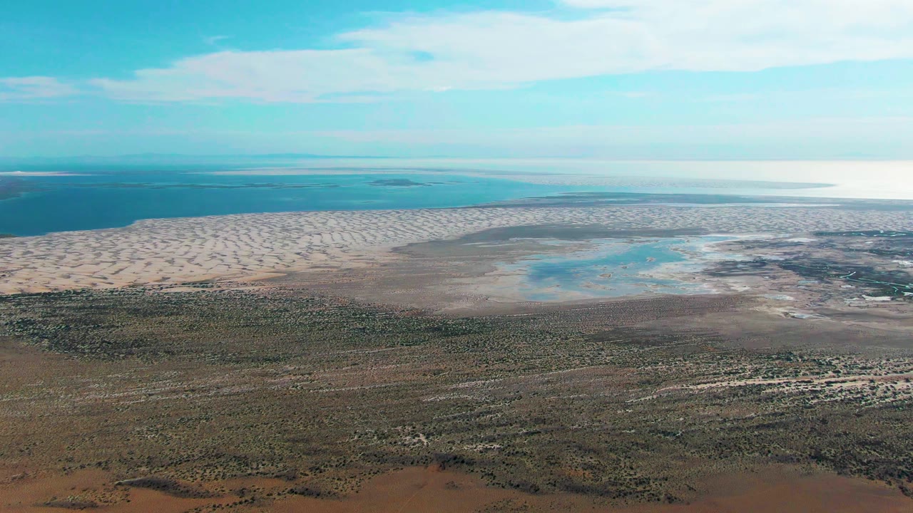 High aerial frame of Guerrero Negro showing brown green scrubland below, pale coastal flats, turquoise shallow pools, deeper water at the horizon, and no visible people.