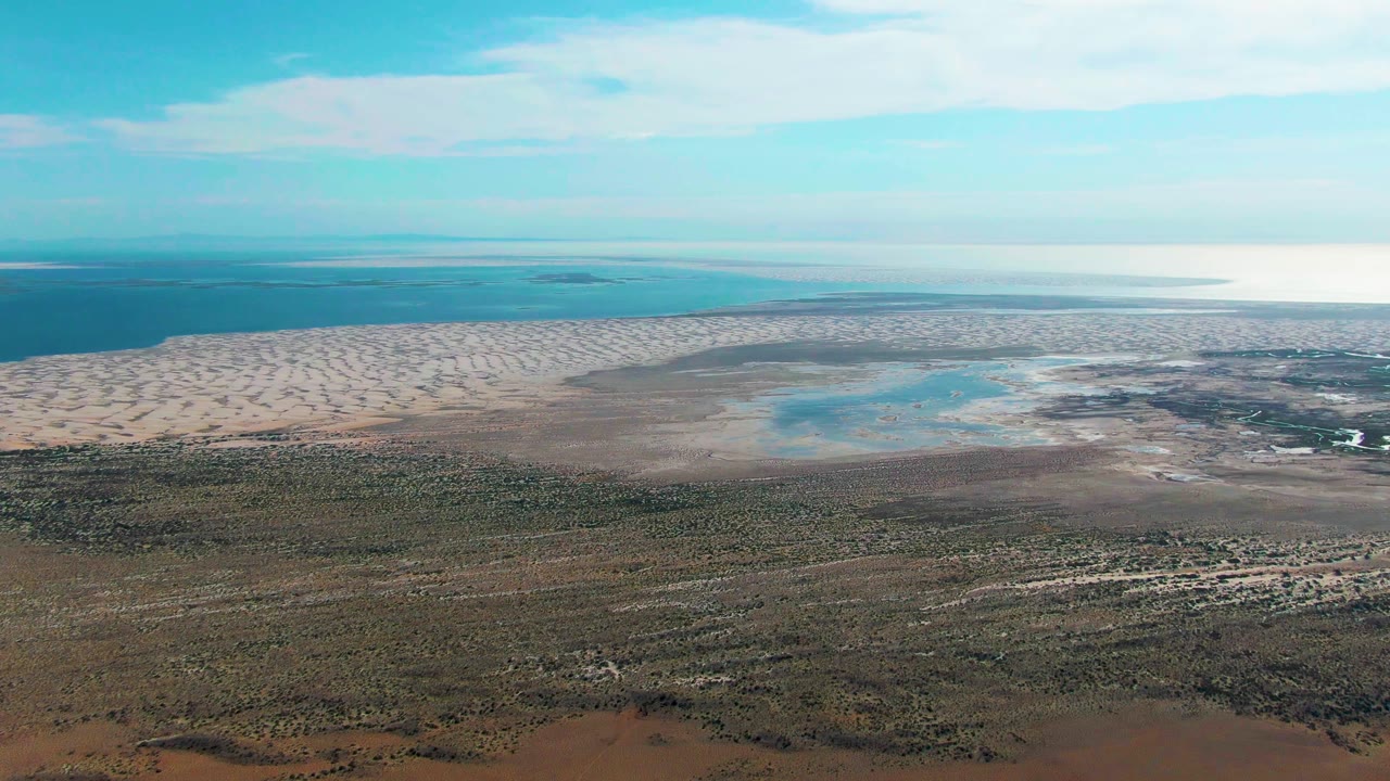 Aerial view in Guerrero Negro with a shallow blue pool set inside pale flats, darker vegetation in the foreground, open water across the distance, and no visible people.