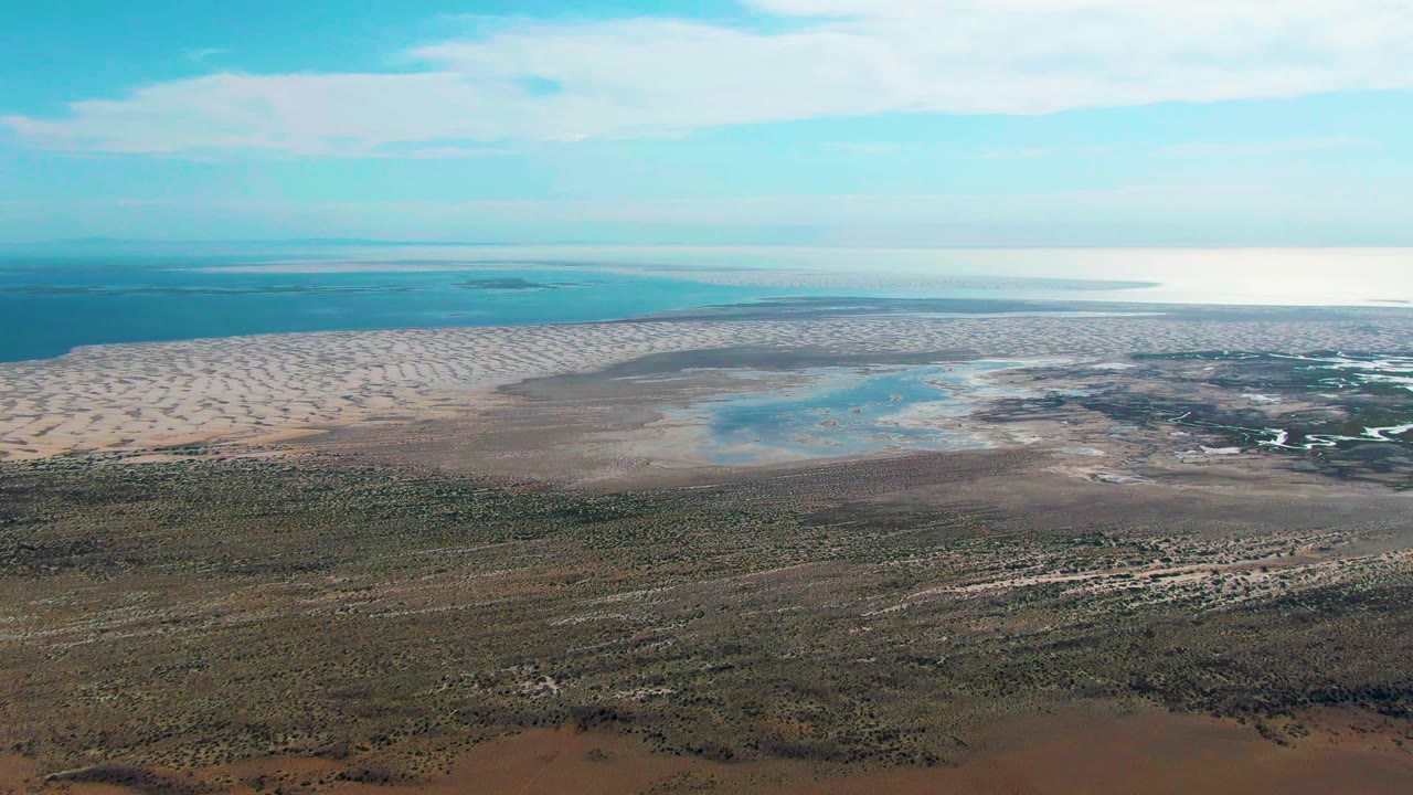 Aerial frame over Guerrero Negro, Baja California Sur showing pale coastal dunes, shallow blue water, foreground scrubland, distant horizon, and no visible people.