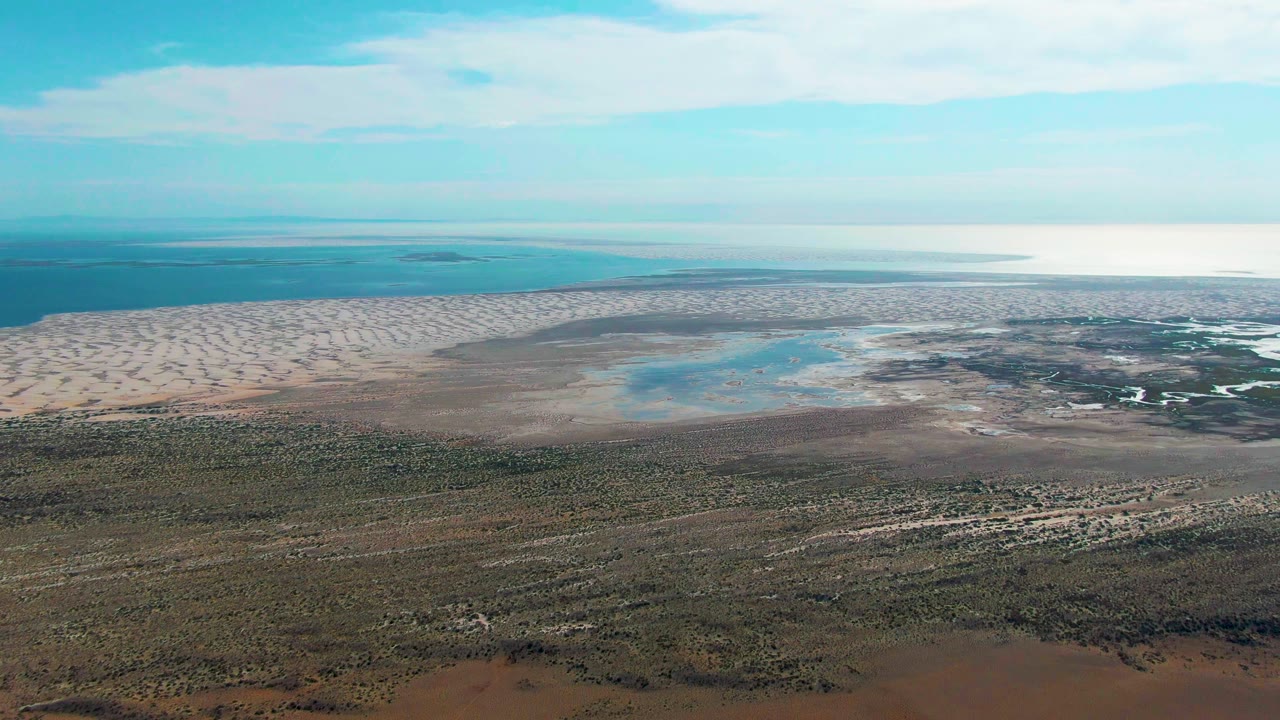 Second aerial frame in Guerrero Negro, Baja California Sur with pale sand bands left of center, shallow water pools in the flats, darker vegetation below, and no visible people.