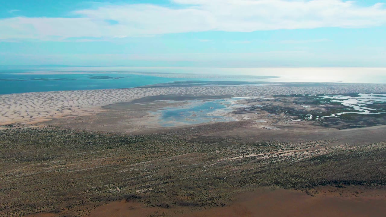 Opening frame over Guerrero Negro tidal flats with scrub in the foreground, shallow blue pools across the flats, a low ocean horizon, partly cloudy sky, and no visible people.