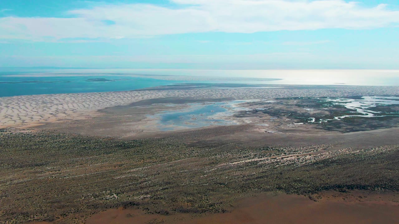 A high view across Guerrero Negro shows pale sand flats on the left, scattered blue water patches near the center, dark marsh channels to the right, and no visible people.