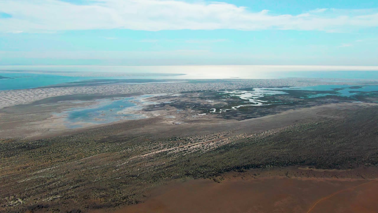 High aerial view over Guerrero Negro coastal wetland, with reddish-brown foreground land, low vegetation, pale flats, turquoise water, a bright horizon, and no visible people.
