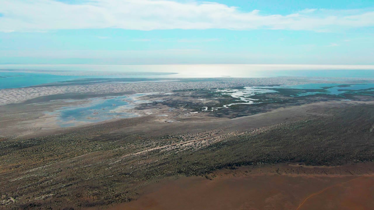 Aerial frame over Guerrero Negro showing low scrubland leading into pale marsh flats, winding water channels, blue-green coastal water, cloud bands, and no visible people.