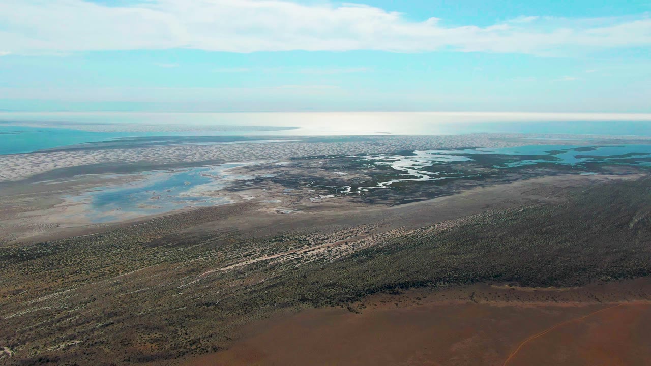 Drone-style aerial over Guerrero Negro with textured brown ground in front, green low vegetation through the center, pale wetland edges, blue shallow water, and no visible people.