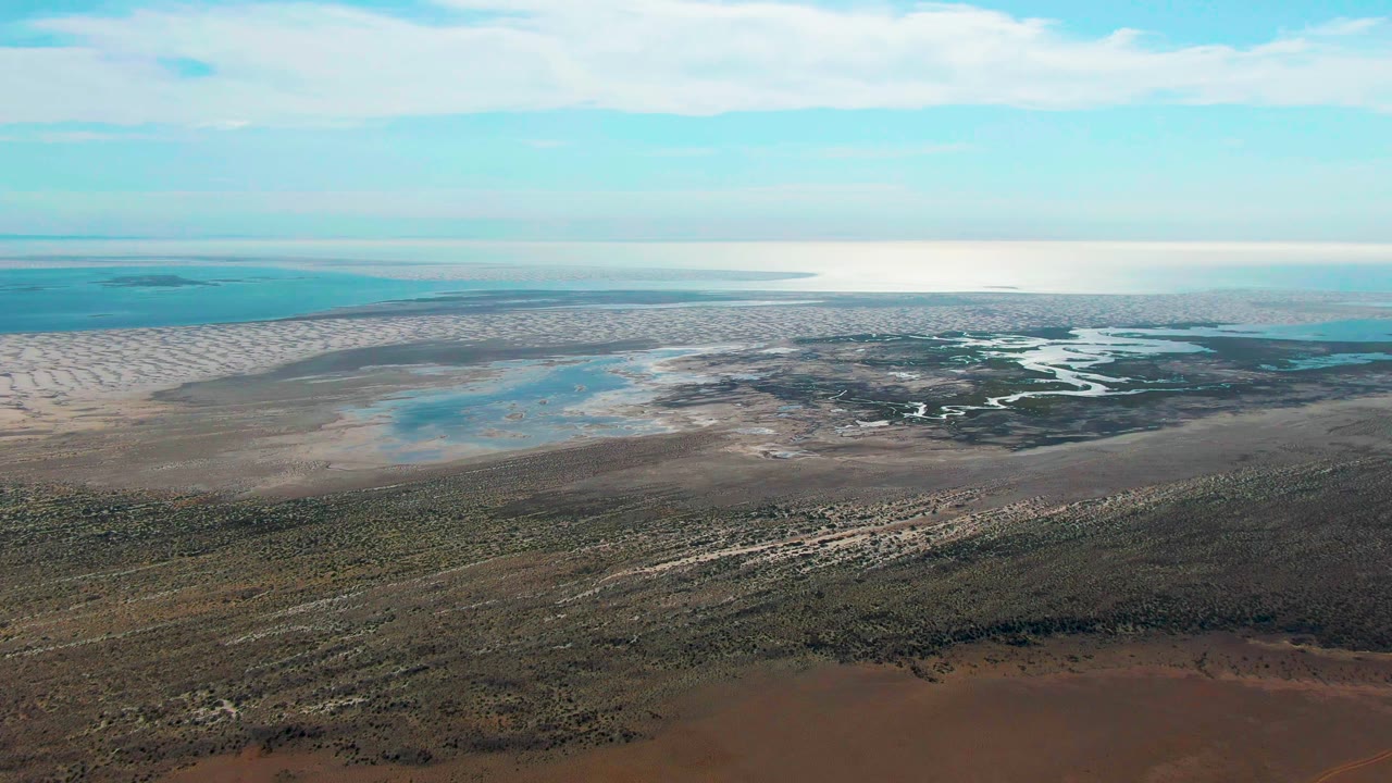 Wide aerial frame over Guerrero Negro, Baja California Sur, with brown scrubland in front, pale wetland flats and blue-green water beyond, and no visible people.