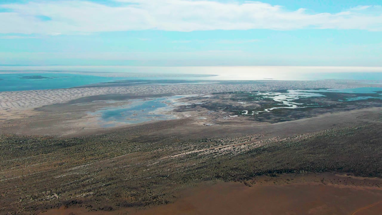 Second aerial frame in Guerrero Negro shows shallow blue water, pale flats, dark wetland channels, brown foreground terrain, and no visible people.