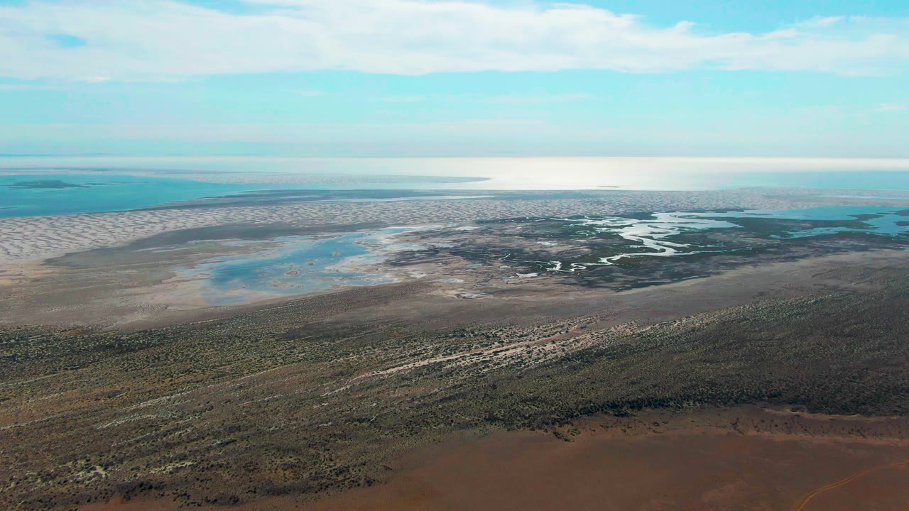 Mid-clip aerial frame from Guerrero Negro with a broad brown foreground, textured vegetation bands, pale wetland flats, blue coastal water, and no visible people.