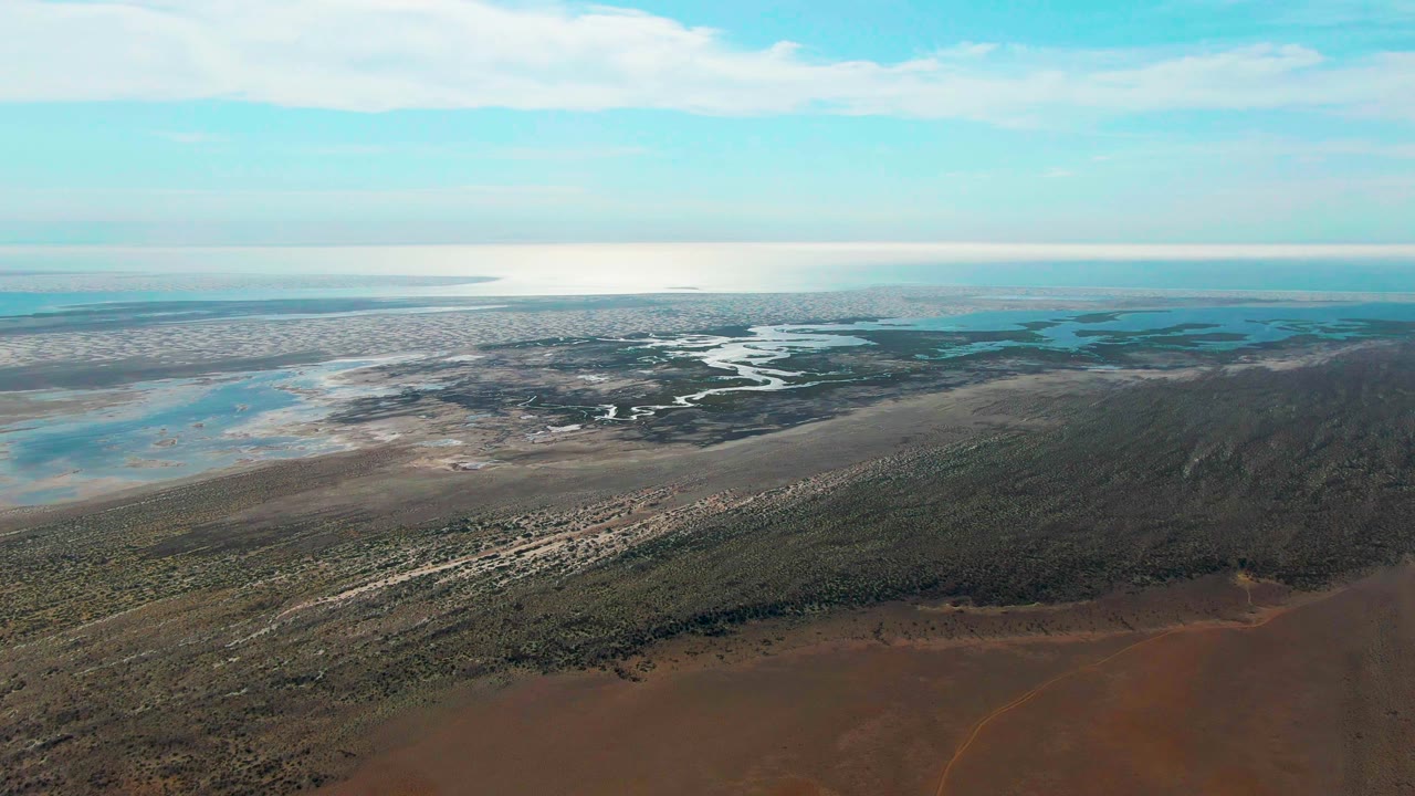 High aerial frame in Guerrero Negro with a red-brown foreground shore, green wetland bands, pale exposed flats, winding water channels, distant coastline, and no visible people.