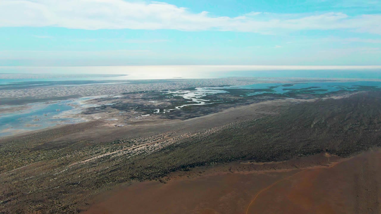 Drone-height view over Guerrero Negro wetlands, with pale mud flats and blue water channels across the background, darker vegetation in the midground, reddish terrain below, and no visible people.