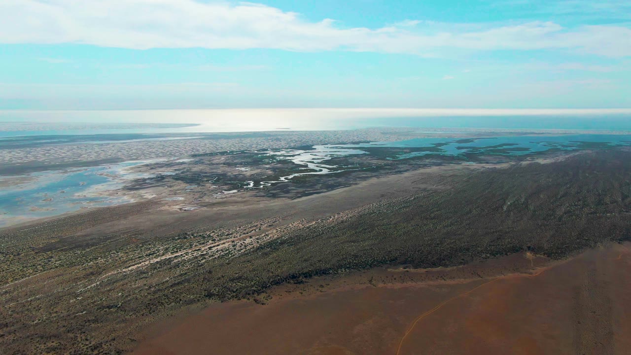 Wide aerial frame in Guerrero Negro showing muddy reddish foreground, a dark green marsh strip, light-toned flats, branching water channels, open water near the horizon, and no visible people.