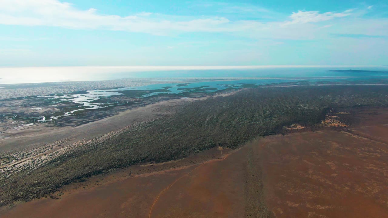 Wide aerial still from Guerrero Negro with a large brown foreground, darker green vegetation across the midground, sandy tidal flats at left, bright water on the horizon, and no visible people.