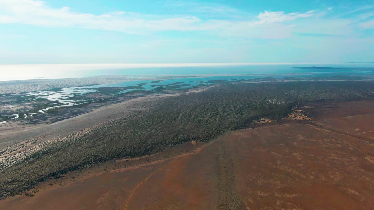 Aerial view of estuary, forest strip, and red dry land under blue sky.