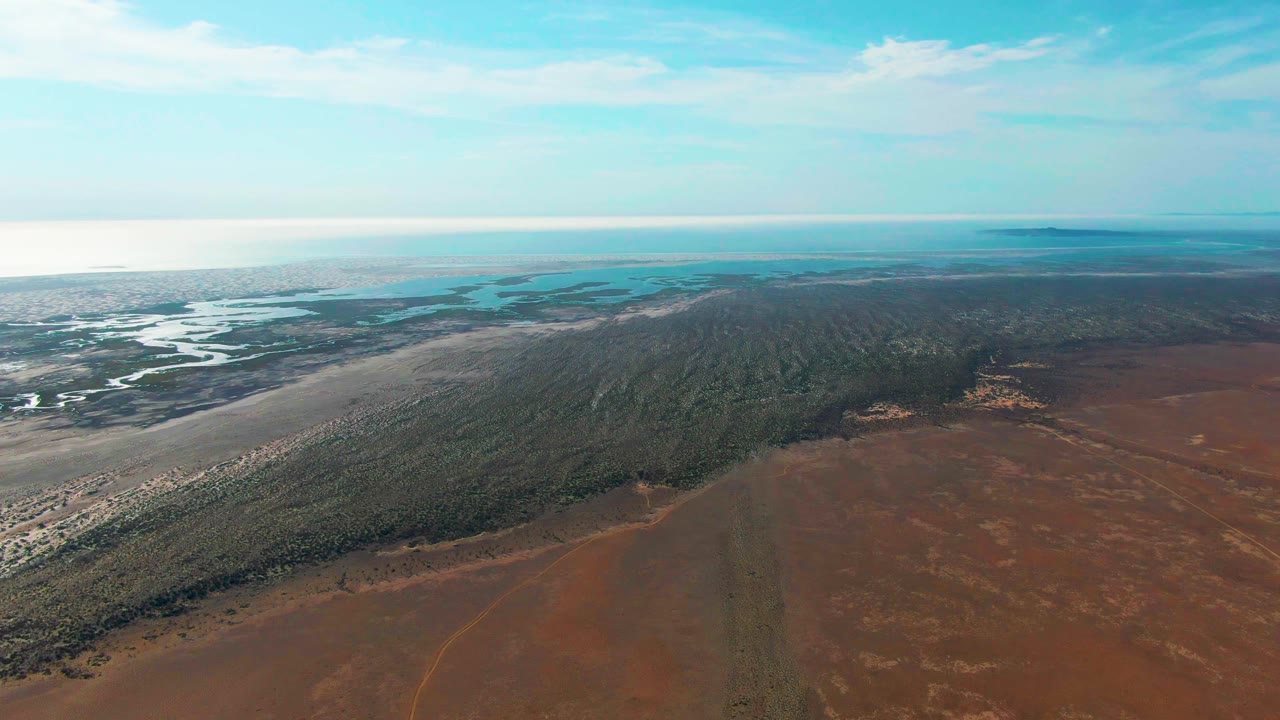 At 0:01, the Guerrero Negro aerial keeps the estuary and pale flats on the left while the dark vegetation band and reddish dry ground occupy more of the lower frame, with no visible people.