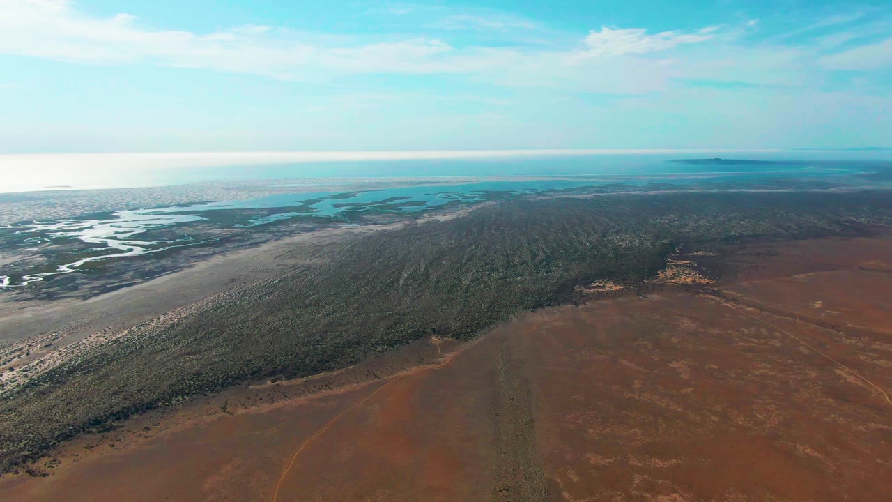 At 0:02, the drone view over Guerrero Negro shows turquoise coastal water beyond pale flats, a continuous dark green strip, rust-colored ground in the foreground, and no visible people.