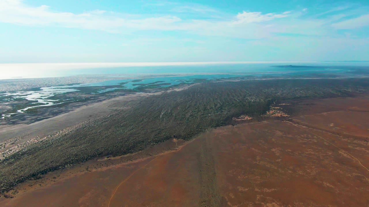 At 0:03, the Guerrero Negro landscape is seen from above with mudflats and water on the left, a forested shoreline across the middle, reddish open land below, and no visible people.