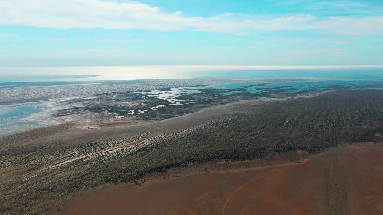 Aerial frame over Guerrero Negro showing brown dry land in front, green wetland vegetation, pale mudflats, winding water channels, ocean on the horizon, blue sky, and no visible people.