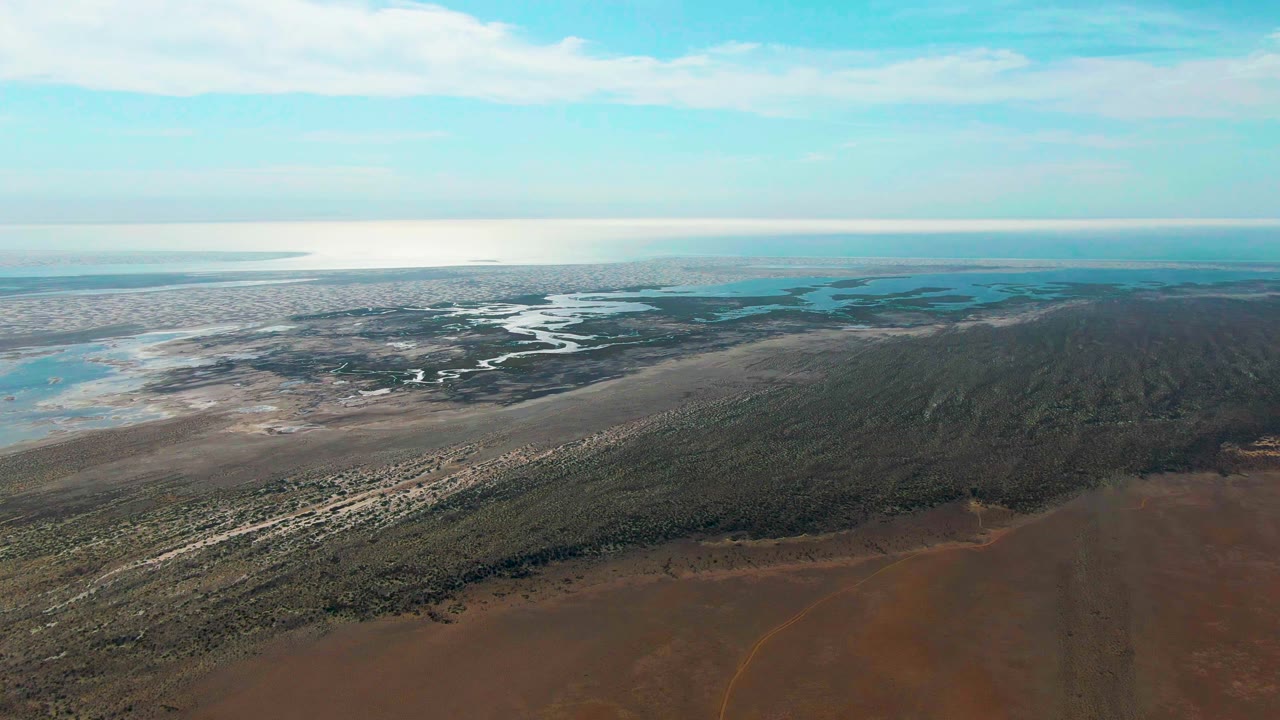 Wide aerial landscape with marsh channels, scrub, mudflats, and distant ocean.