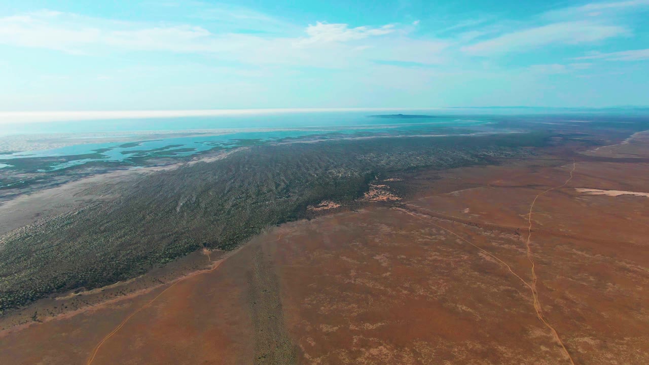 Wide aerial view over Guerrero Negro, Baja California Sur, with reddish dry field below, a dark forest band through the middle, distant pale water near the horizon, and no visible people.