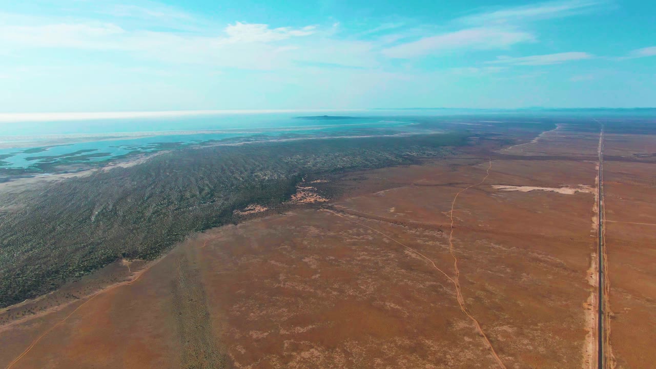At 0:02, the Guerrero Negro landscape shows a stronger view of the straight road along the right edge, open brown terrain across the foreground, forest stretching left of center, distant light blue water or flats, and no visible people.
