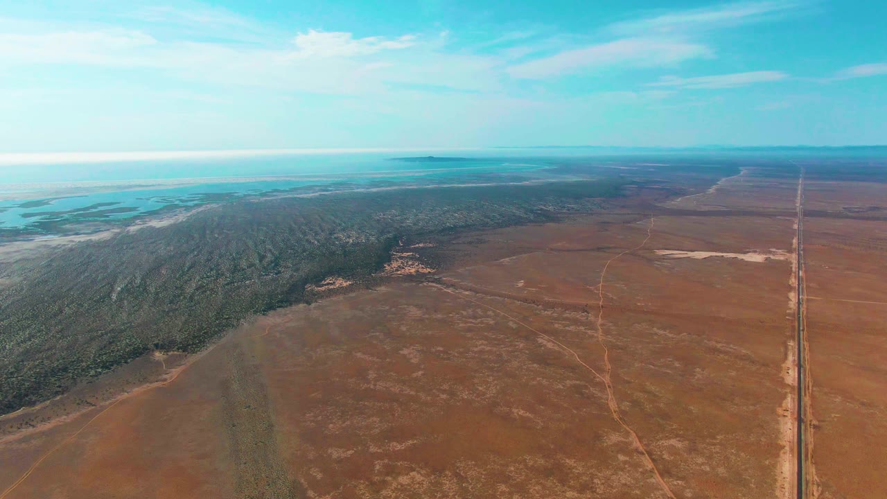 At 0:03, the Guerrero Negro aerial composition centers the forest edge and brown open land, with the road still near the right border, pale water or flats along the far left horizon, soft clouds overhead, and no visible people.