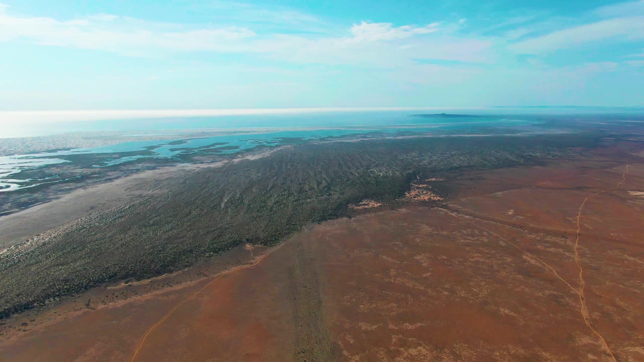 Second aerial frame in Guerrero Negro showing more of the brown open plain below the forest edge, with turquoise water and a bright clouded sky beyond, and no visible people.