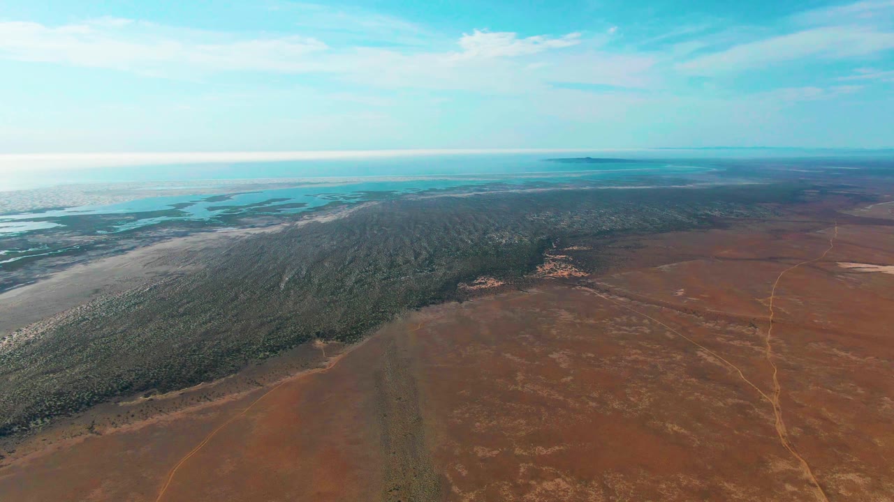 Wide aerial of dry land beside forest with water on the horizon.