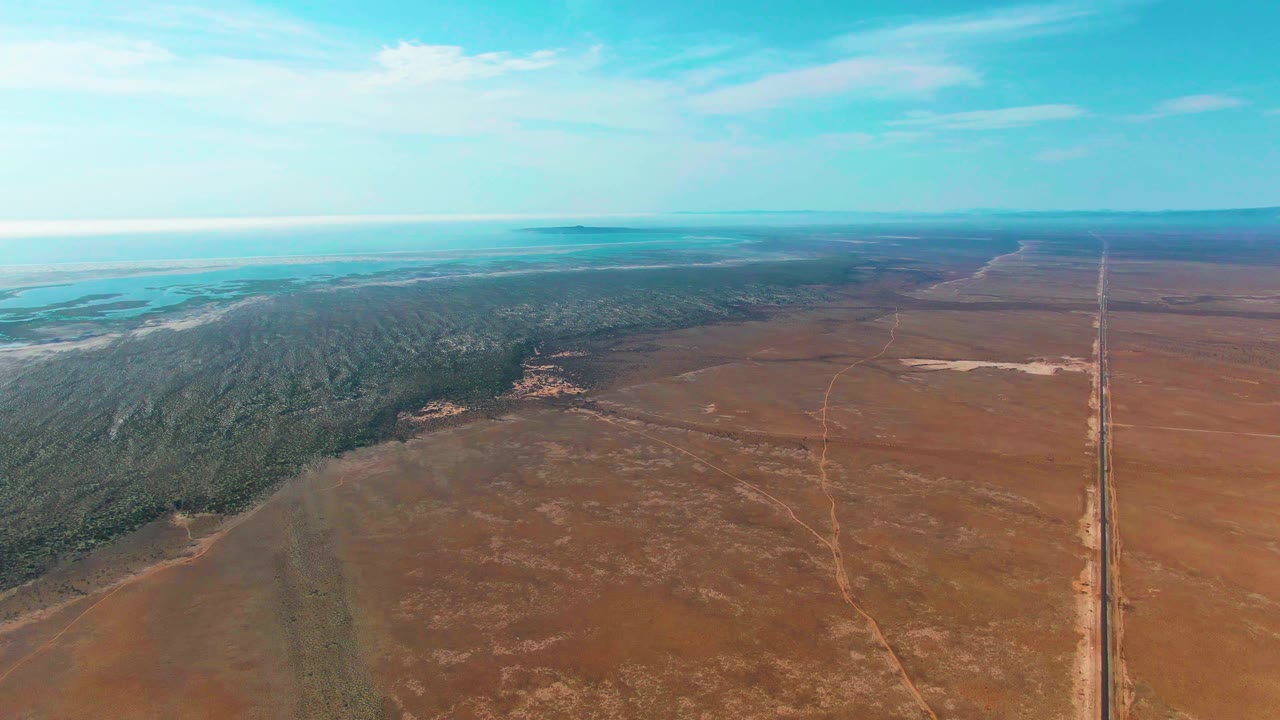 Opening aerial frame over Guerrero Negro, Baja California Sur, with dry brown fields in front, a green forested band to the left, a straight road to the right, distant pale water, and no visible people.