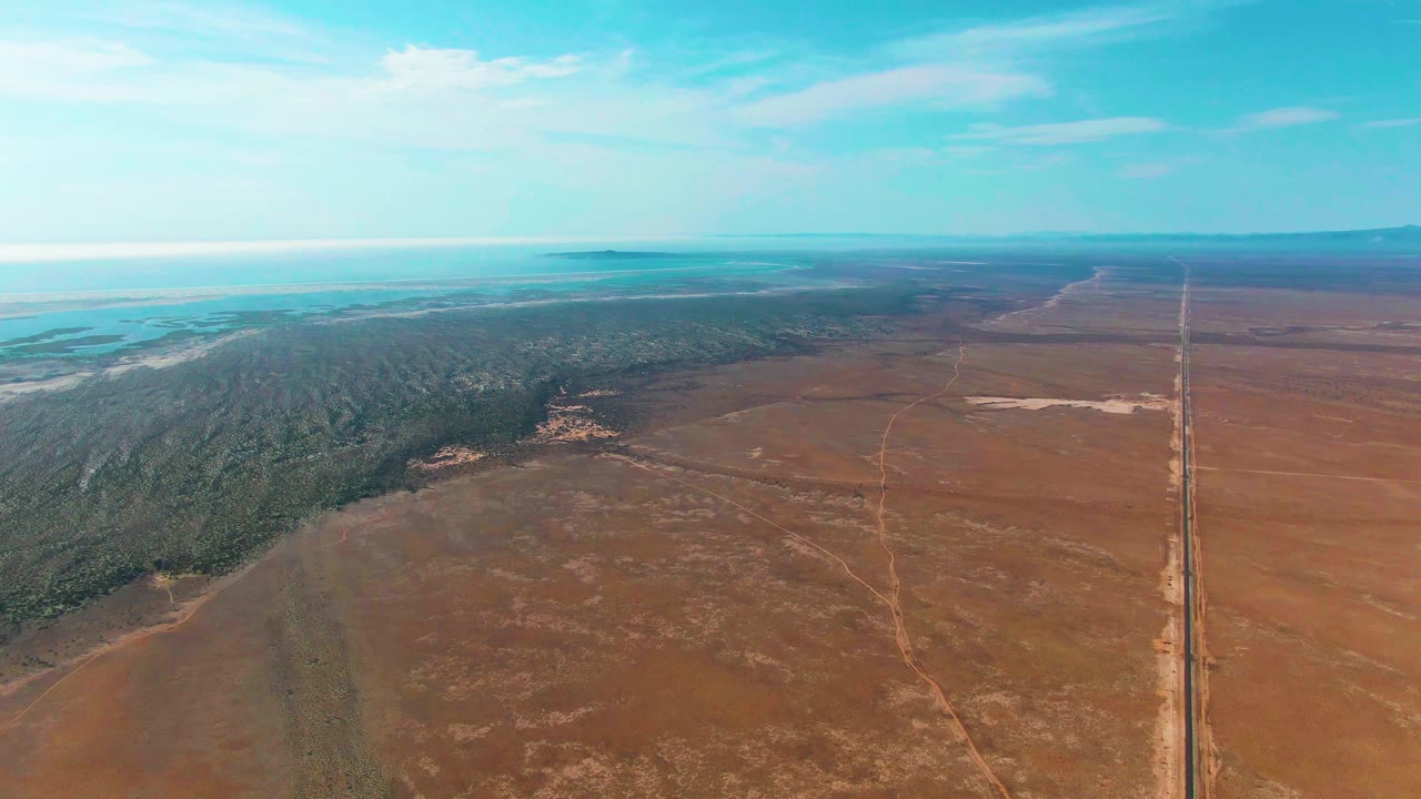 High aerial view in Guerrero Negro with dry fields dominating the foreground, a dark green forested edge left of center, a narrow straight road on the right, bright distant flats or water, and no visible people.