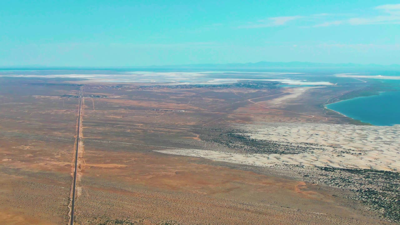 Opening aerial frame over Guerrero Negro, Baja California Sur, with a straight road running through brown desert plains toward white salt flats and blue water, no visible people.