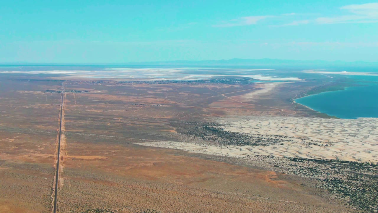 Third frame in Guerrero Negro keeps the long road prominent at left, with brown open plain, white salt deposits, distant horizon, blue sky, and no visible people.