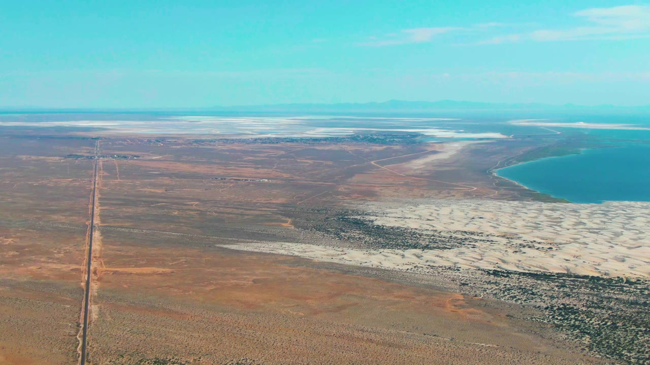 Straight road and desert plain seen from above, with white salt flats and blue water nearby.