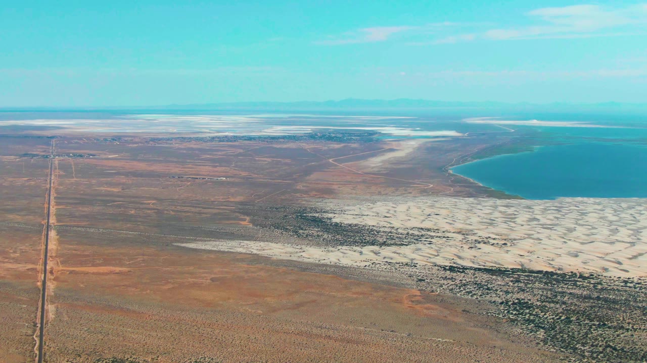 Aerial view in Guerrero Negro with the vertical road slightly left of center, brown desert flats, a broad white salt-flat band, turquoise lake water on the right, low horizon, and no visible people.