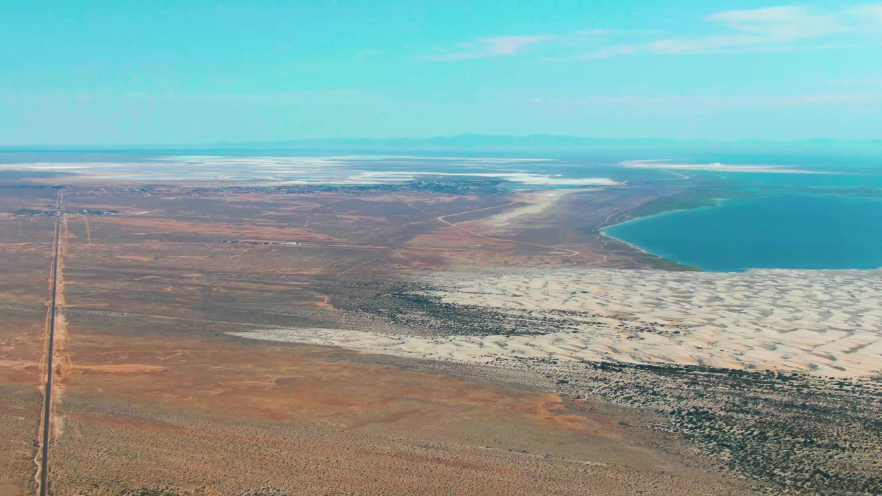 High aerial frame over Guerrero Negro desert terrain, with a straight road running through the left side, white salt flats spreading across the lower right, blue lake water beyond them, mountains on the horizon, and no visible people.