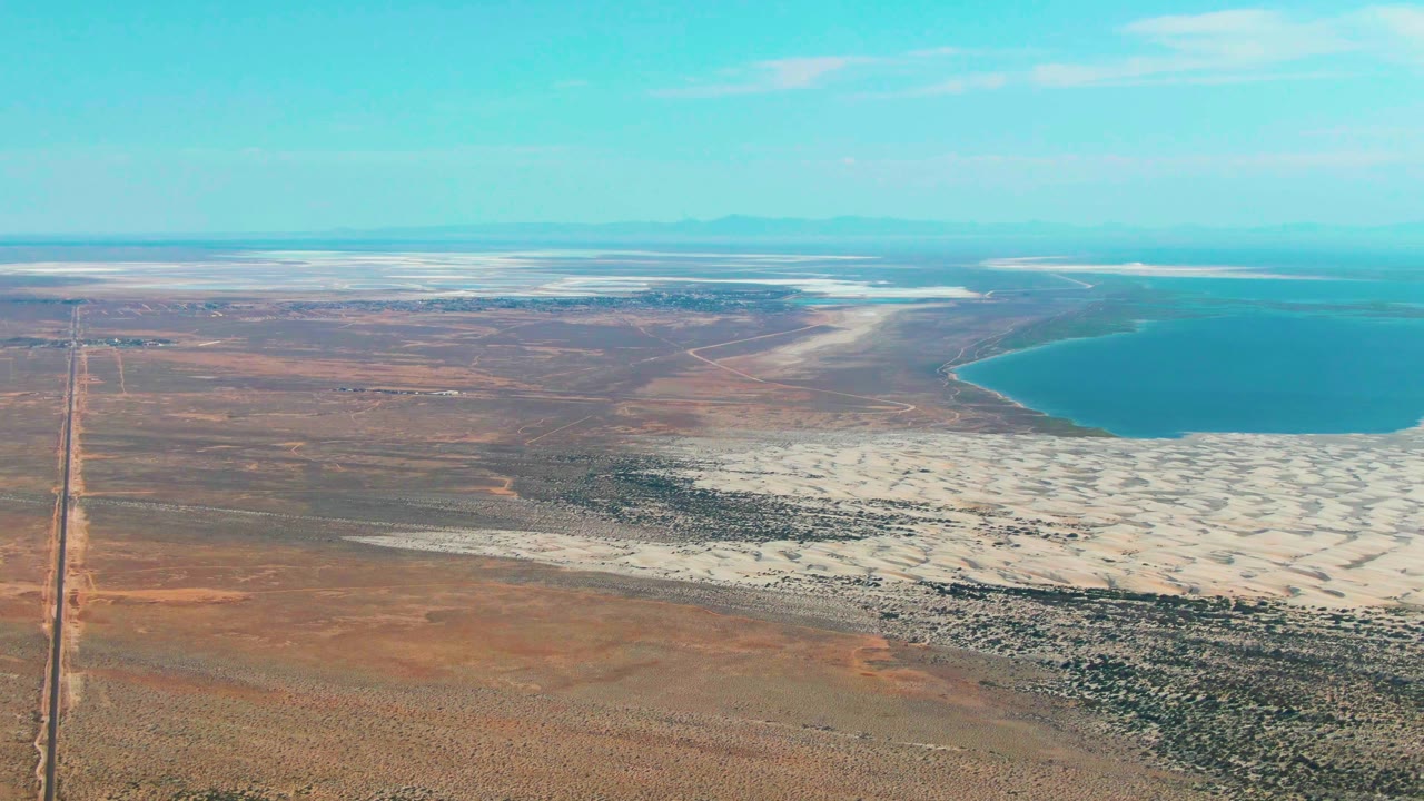 Guerrero Negro aerial frame with the road along the left edge, reddish-brown desert in the lower foreground, white salt deposits across the middle right, blue lake water at upper right, a flat horizon, and no visible people.