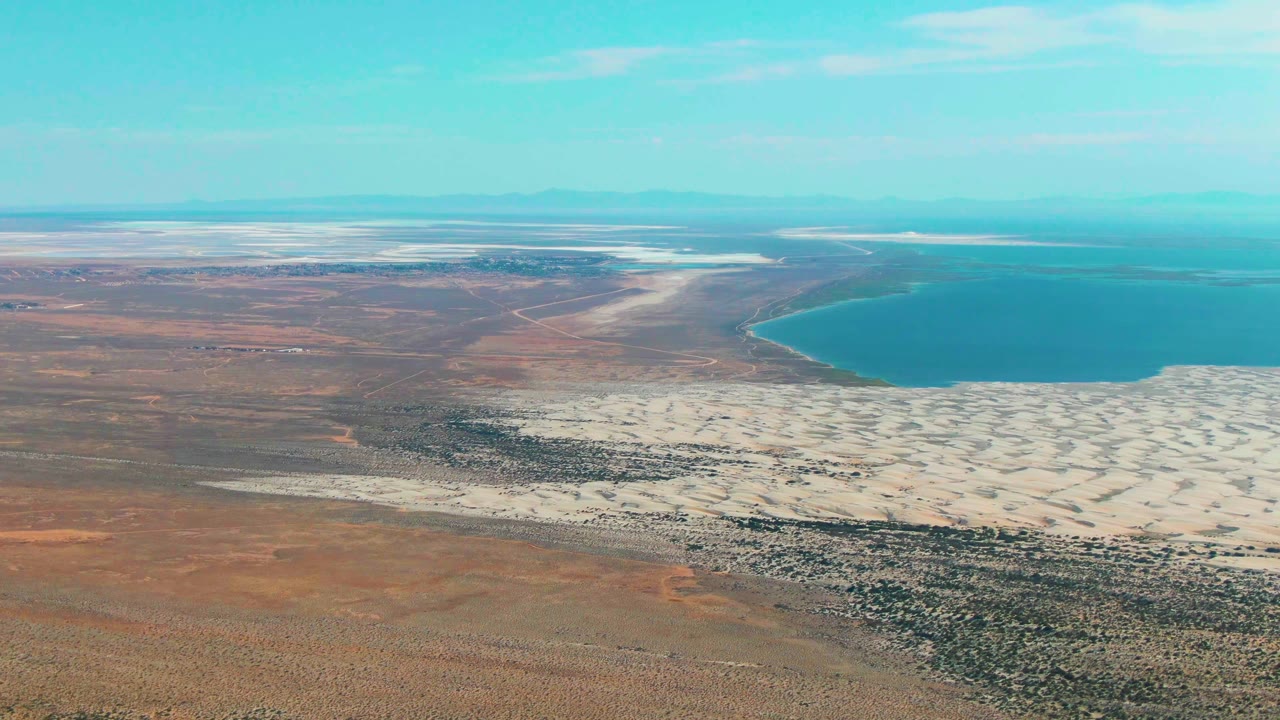 Aerial desert plain, white salt flat, and blue water under a pale sky.
