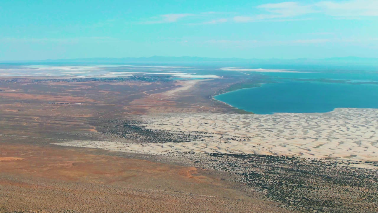Aerial frame over Guerrero Negro showing more of the white salt flat across the midground, rust-brown land in the foreground, calm blue water in the background, and no visible people.