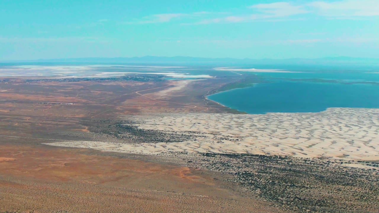 Wide aerial view in Guerrero Negro with the shoreline curving around blue water, pale salt flats across the center, dry brown land below, and no visible people.
