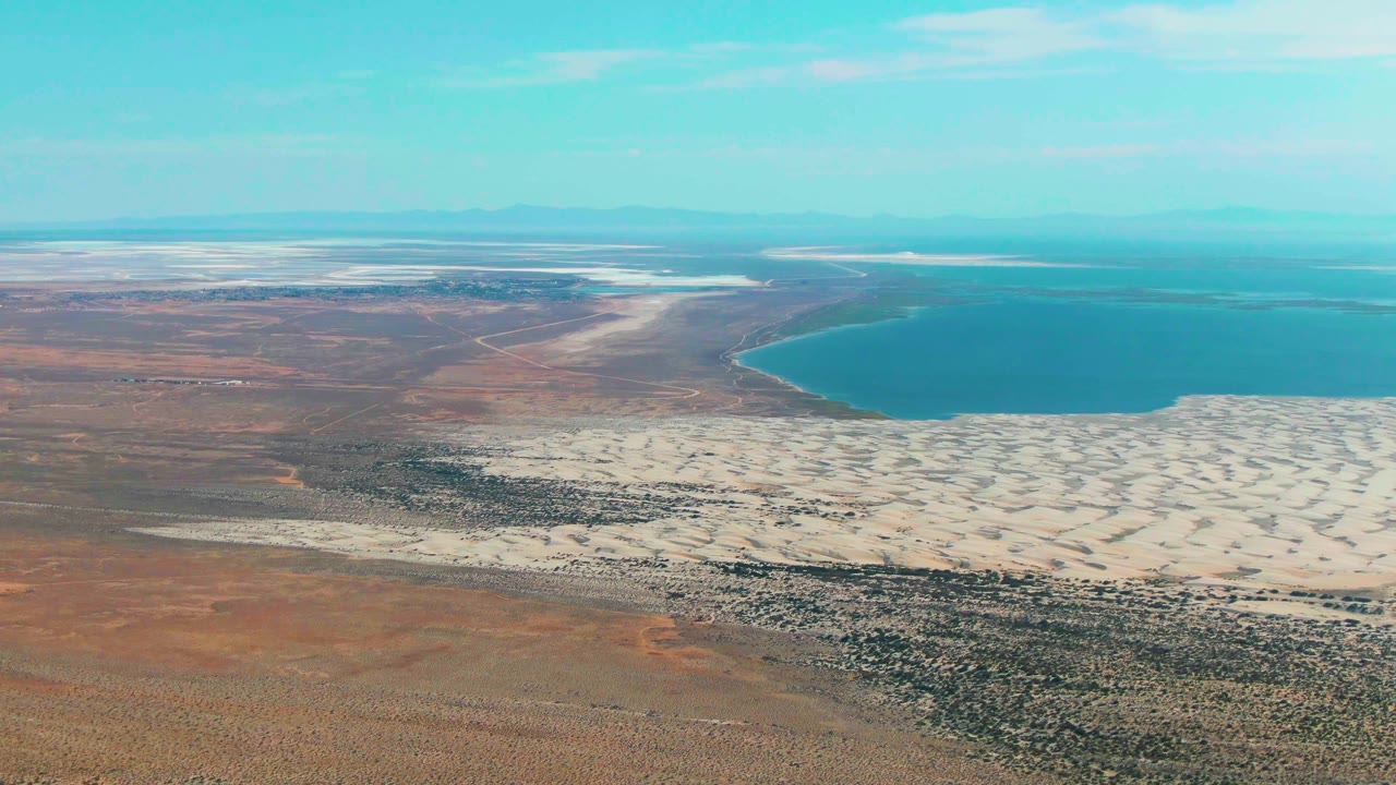 Mid-clip aerial frame over Guerrero Negro showing brown desert foreground, pale salt flat near the water, blue bay-like water, distant low mountains, and no visible people.
