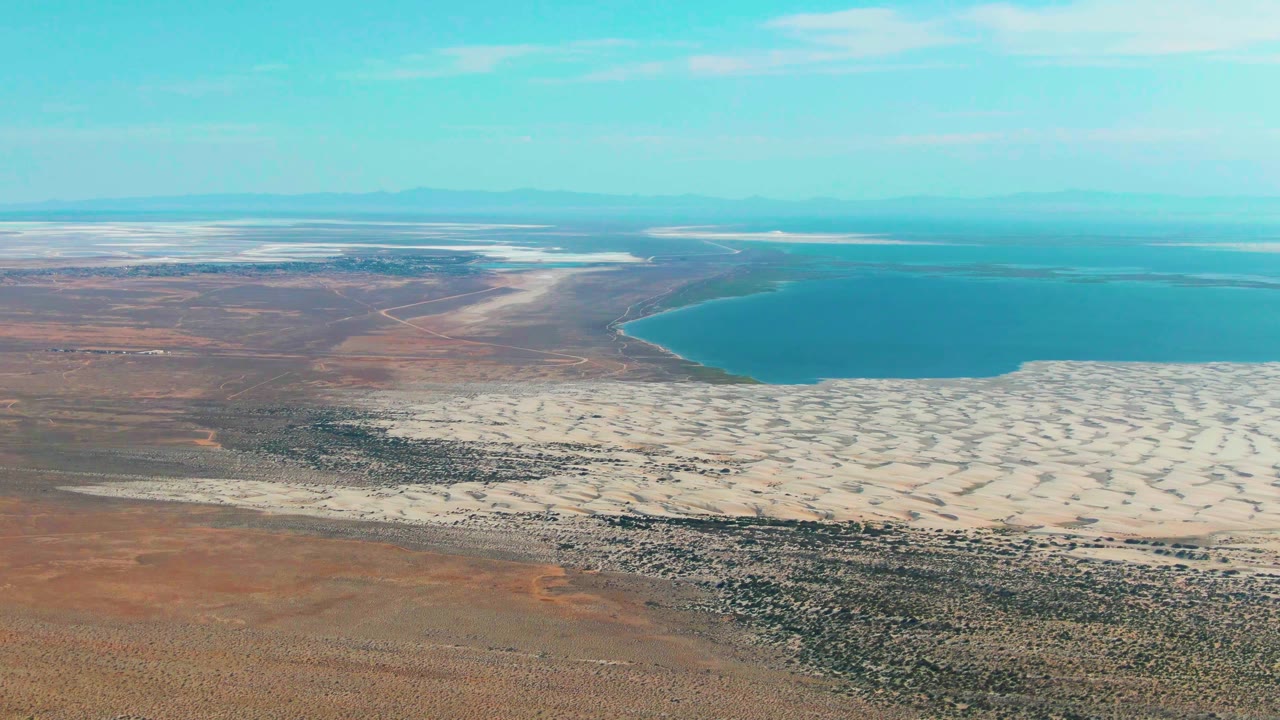 Opening aerial view over Guerrero Negro, Baja California Sur, with reddish desert foreground, pale dune fields, a broad blue lake, distant ridges, and no visible people.