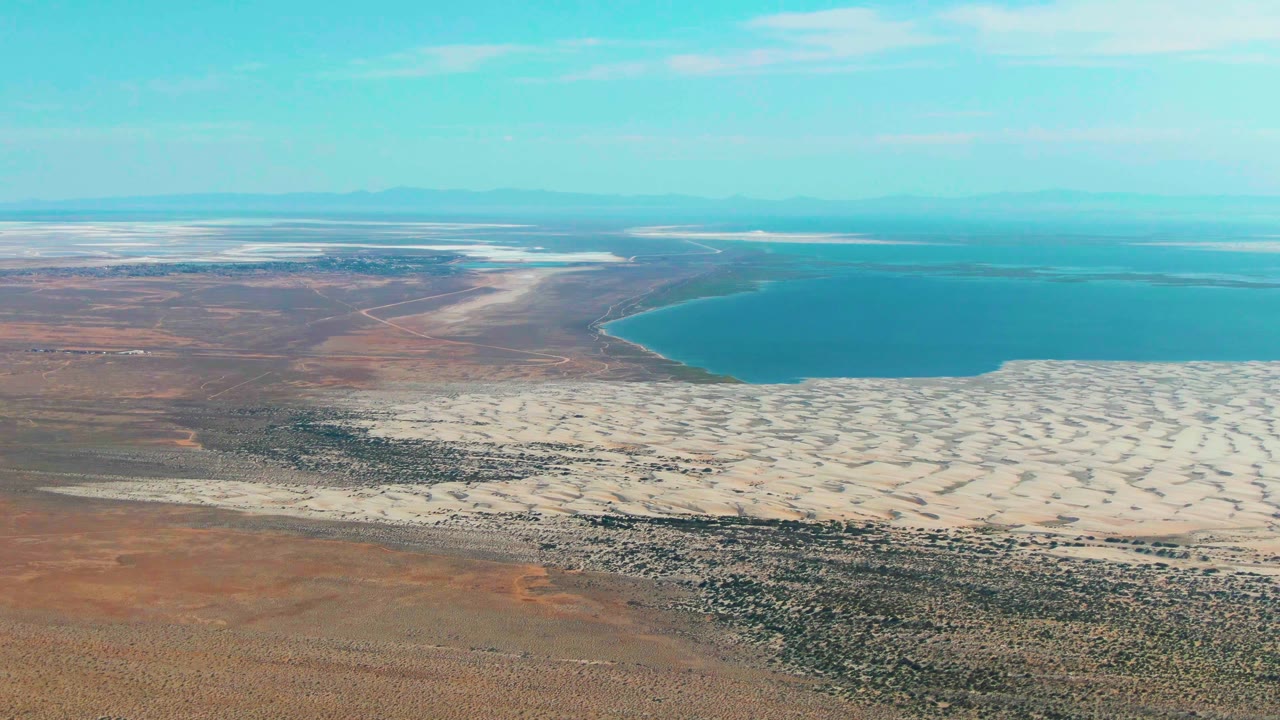 Aerial frame from Guerrero Negro shows pale dune patterns spreading below a curved blue shoreline, with dry brown terrain to the left and no visible people.
