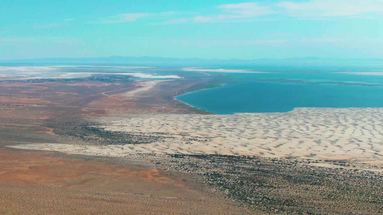 The aerial view holds on the Guerrero Negro lake edge, with pale dune ridges across the lower center, reddish arid ground at left, hazy mountains beyond, and no visible people.