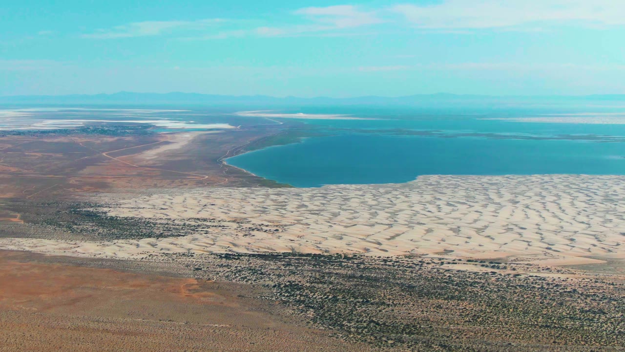 Aerial frame over Guerrero Negro, Baja California Sur showing scrub desert and pale dunes leading to a blue lake, with distant flats on the horizon and no visible people.