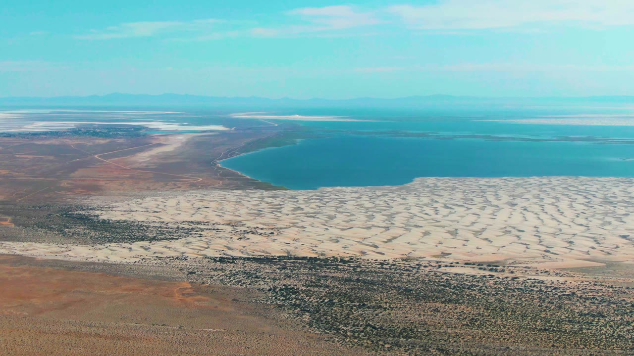 Wide aerial frame in Guerrero Negro, Baja California Sur with brown desert foreground, bright dune bands, a curved blue lake edge, distant flats, and no visible people.