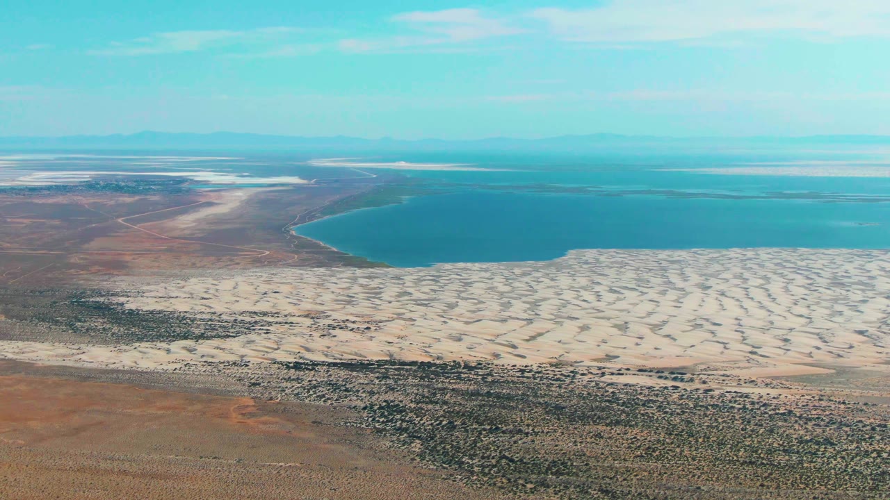 High aerial frame over Guerrero Negro, Baja California Sur showing textured scrubland below, light sand dunes across the middle, blue lake water beyond, and no visible people.