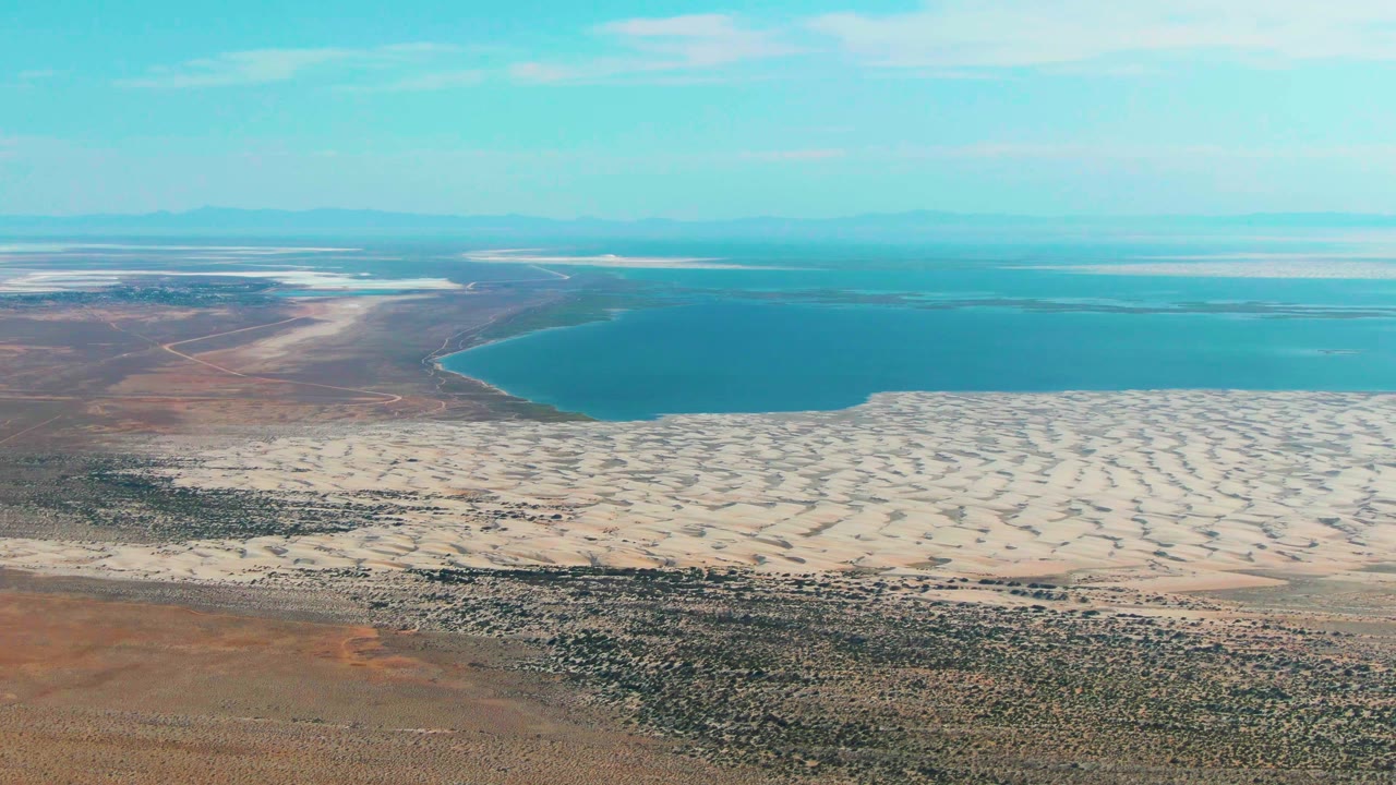 Desert foreground and pale dunes border a blue lake beneath a bright cyan sky.