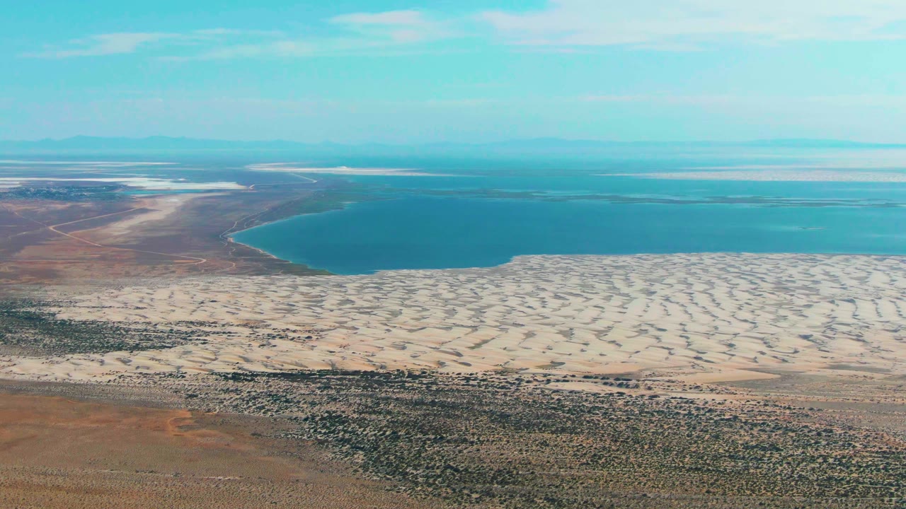 Aerial frame in Guerrero Negro shows the curved edge of blue water pressing against pale sand, with brown arid land and scattered vegetation below, no visible people.