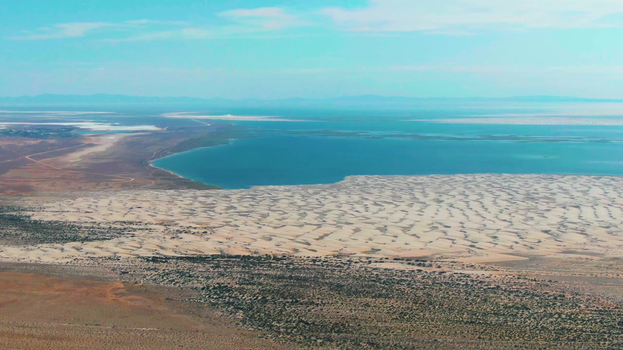 Wide aerial view in Guerrero Negro with turquoise water across the upper frame, rippled pale sand through the center, and scrubby desert foreground, no visible people.